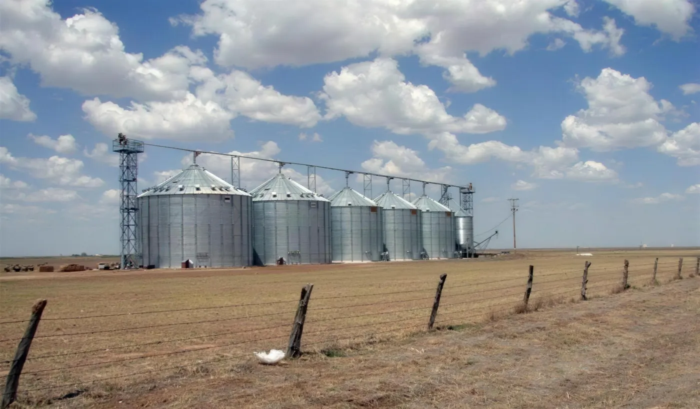 Six steel grain silos on flat Panhandle farmland, with a barbed-wire fence in front and clouds above.