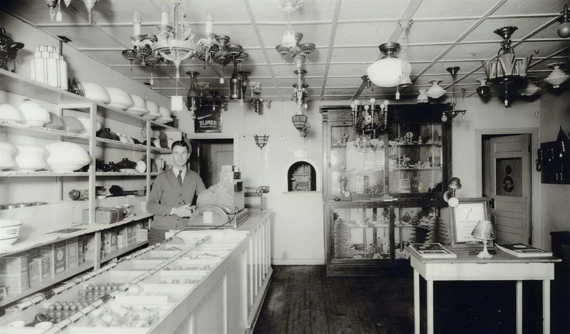 Black and white photo of a vintage shop interior from Triangle Electric's website: a man in a cardigan stands behind a counter, with glass light shades on shelves and chandeliers hanging from the ceiling.