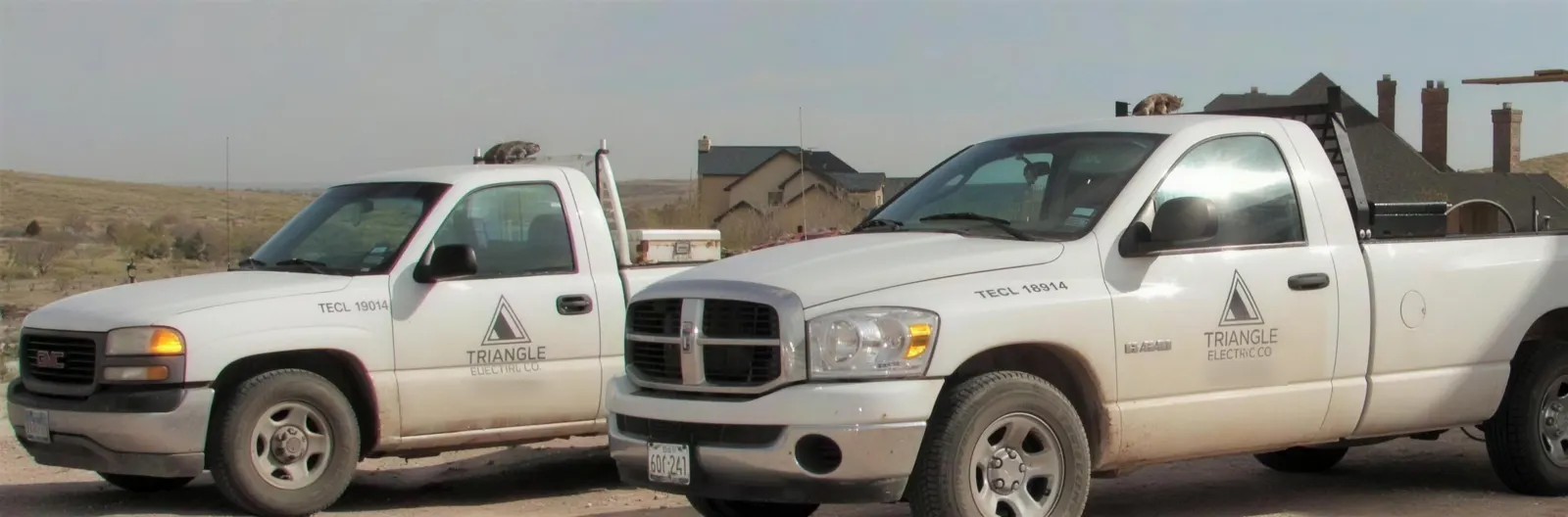 Two white Triangle Electric Co. pickup trucks parked on a dirt lot in front of a new house.
